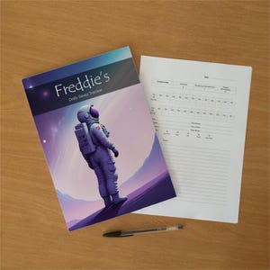 Könnte beinhalten: Ein tägliches Schlaf-Tagebuch mit dem Titel Freddie's Daily Sleep Tracker, das einen Astronauten in einem violetten Raumanzug auf dem Einband zeigt, zusammen mit einem weißen Datenblatt und einem schwarzen Kugelschreiber zur Schlafaufzeichnung.
