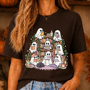 May include: A dark brown t-shirt featuring a graphic print of seven white ghosts reading books on a wooden bookshelf. The design includes green vines, lit candles, a small black cat, and stacks of colourful books surrounding the ghosts.