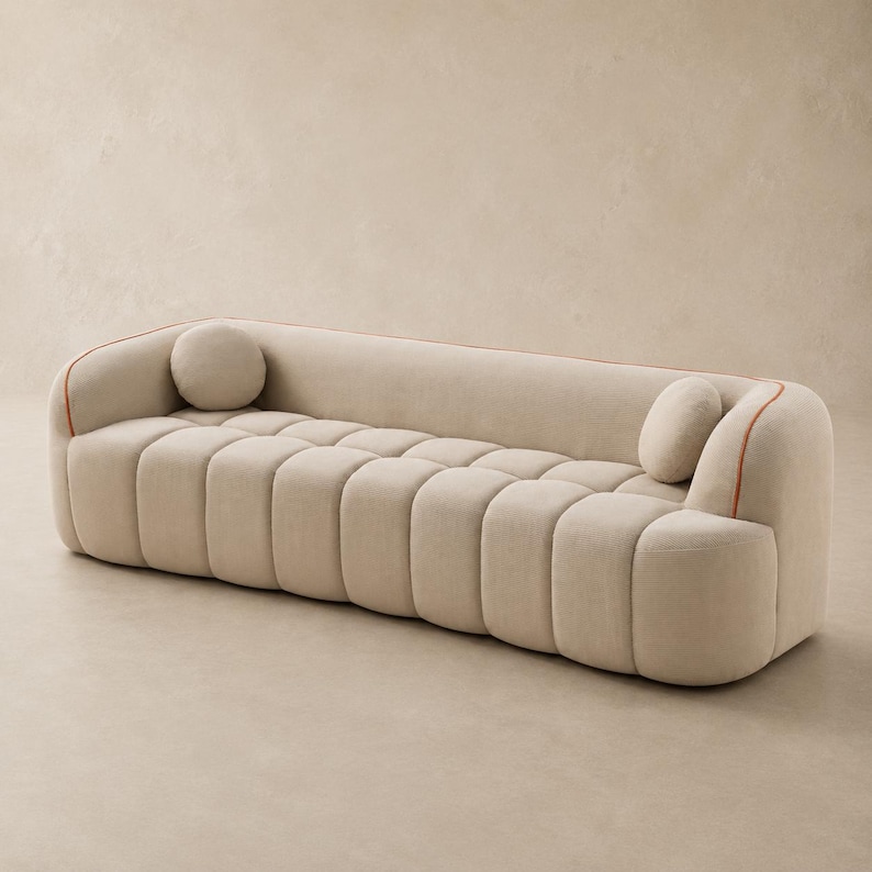 May include: A beige sofa with a channel-tufted seat and backrest, featuring rounded armrests and a thin orange piping detail along the top edges. The piece includes two small, round, matching beige accent cushions placed at each end.