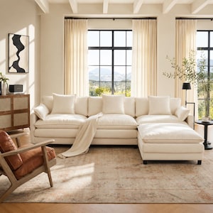 May include: A cream-colored sectional sofa with multiple throw pillows, a matching ottoman, and a light-colored throw blanket. The living room features a patterned rug, a wooden chair with a leather seat, and a cabinet with a black and white abstract painting.