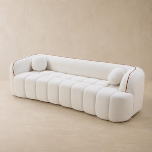 May include: A white sofa with a channel-tufted seat and backrest, featuring rounded edges and a brown piping detail along the top frame. Two small, round, matching white throw pillows are positioned at each end of the seating area.