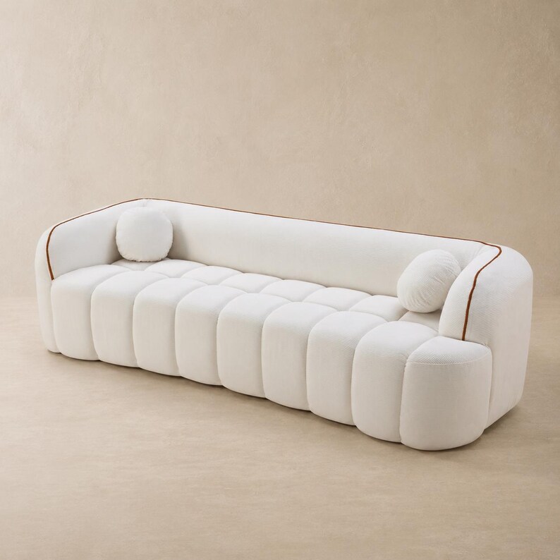 May include: A white sofa with a channel-tufted seat and backrest, featuring rounded edges and a brown piping detail along the top frame. Two small, round, matching white throw pillows are positioned at each end of the seating area.