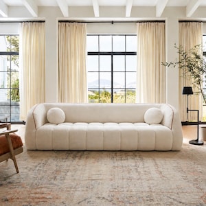 May include: A cream-colored, low-profile sofa with a channel-tufted seat and backrest. The sofa features rounded arms, a thin brown piping detail along the edges, and two matching round throw pillows placed at each end of the seating area.