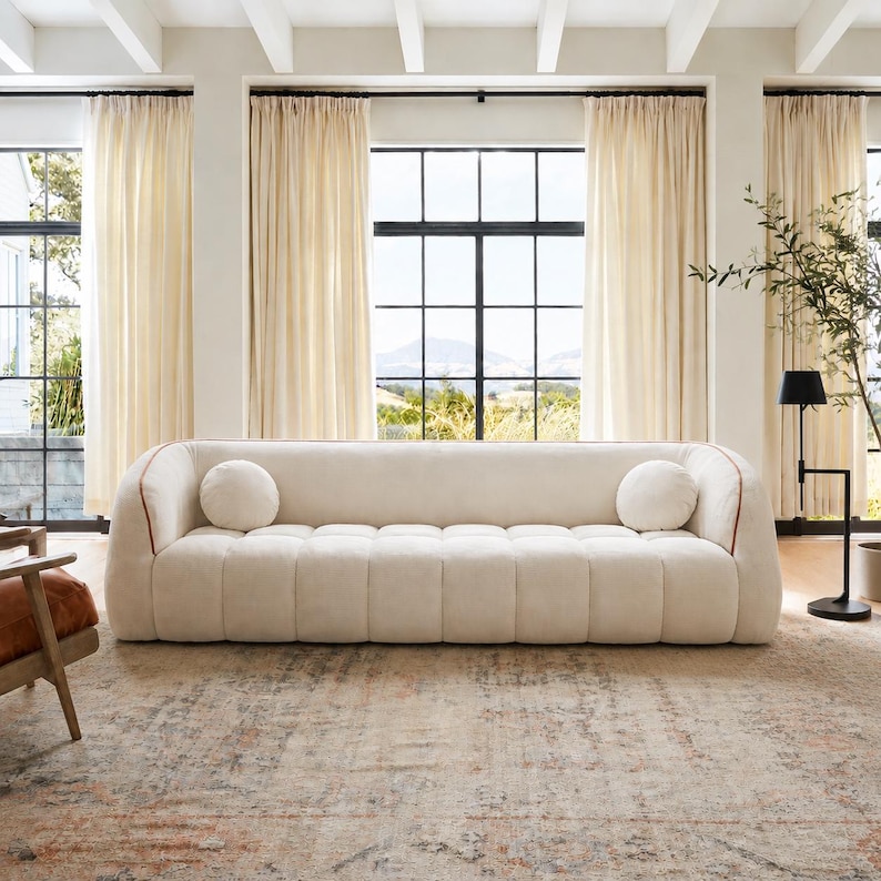 May include: A cream-colored, low-profile sofa with a channel-tufted seat and backrest. The sofa features rounded arms, a thin brown piping detail along the edges, and two matching round throw pillows placed at each end of the seating area.