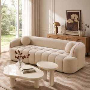 May include: A cream-colored sofa with a channel-tufted seat and back, featuring rounded arms, two matching round throw pillows, and thin orange piping accents. The sofa is paired with two glossy, off-white, organic-shaped coffee tables.
