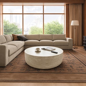 May include: A modern living room featuring a large, beige sectional sofa, a round stone coffee table, and a patterned rug. A cup of coffee, a book, and a remote are on the table. A floor lamp and large windows with a city view complete the scene.