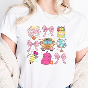 Op de afbeelding: Een wit t-shirt met een kleurrijke grafische print van schoolgerelateerde items, waaronder een stapel boeken, roze strikken, een doos kleurpotloden, een gele schoolbus, een wereldbol, een potlood, een rugzak en kleine ster- en bloemaccenten.