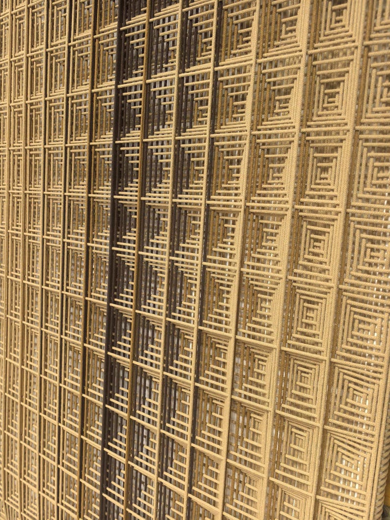 Puede incluir: Un tapiz de pared con un patr&oacute;n de cuadr&iacute;cula geom&eacute;trica compuesto por cuerdas de fibra en capas con forma cuadrada. El dise&ntilde;o muestra un degradado de tonos neutros, desde beige claro y tostado hasta marr&oacute;n oscuro, creando un motivo texturizado.