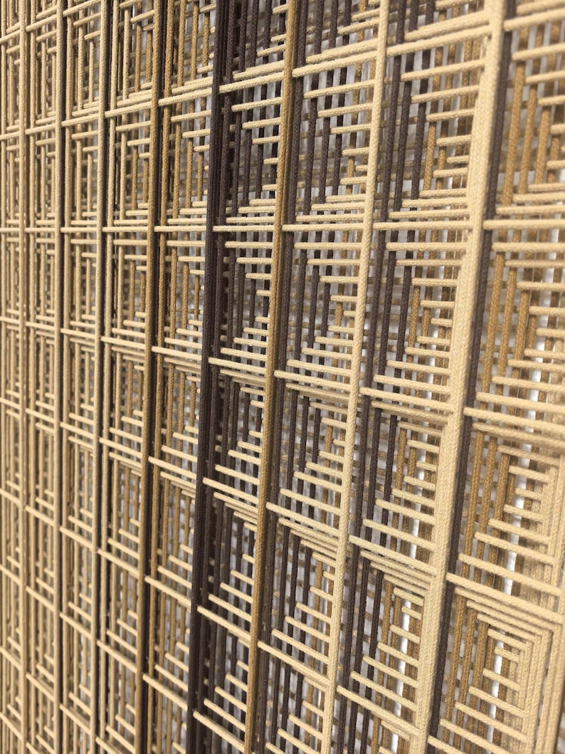 Puede incluir: Una pieza de arte mural que presenta una serie de paneles verticales compuestos por hilos estrechamente tejidos en tonos beige, bronceado y marr&oacute;n oscuro. Los hilos est&aacute;n dispuestos en un patr&oacute;n de rejilla geom&eacute;trica repetitivo.