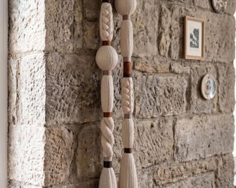 Puede incluir: Un tapiz de pared que presenta dos largas hebras verticales de fibra suspendidas de un marco rectangular de madera clara. Las hebras consisten en secciones de hilo color crema, elementos trenzados, nudos esféricos y acentos marrón oscuro, terminando en borlas.