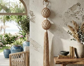 Puede incluir: Una decoración de pared de macramé con dos formas esféricas envueltas en una red de cuerda anudada de color tostado, suspendida de un gancho de metal color latón. La pieza incluye un largo fleco de cuerda tostada adornado con pequeñas cuentas de madera.