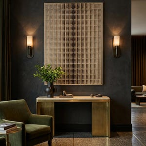 Puede incluir: Una pieza de arte mural rectangular con un patr&oacute;n de cuadr&iacute;cula en tonos beige y marr&oacute;n oscuro, montada sobre una mesa consola de lat&oacute;n con una superficie de piedra clara. Un jarr&oacute;n negro con hojas verdes descansa sobre la mesa, flanqueado por dos apliques de pared con pantallas blancas.