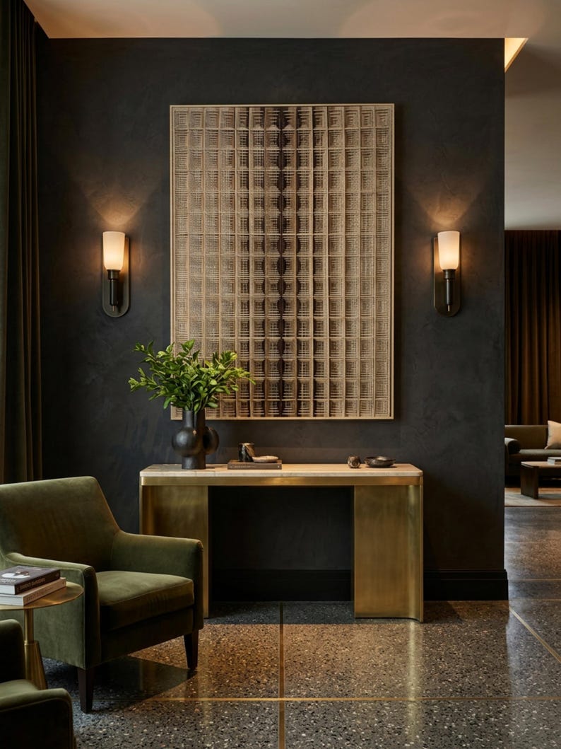 Puede incluir: Una pieza de arte mural rectangular con un patr&oacute;n de cuadr&iacute;cula en tonos beige y marr&oacute;n oscuro, montada sobre una mesa consola de lat&oacute;n con una superficie de piedra clara. Un jarr&oacute;n negro con hojas verdes descansa sobre la mesa, flanqueado por dos apliques de pared con pantallas blancas.