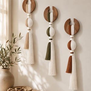 Op de afbeelding: Drie wandhangers met halvemaanvormige donkerhouten bovenkanten en verticale vezelkwasten in wit gecombineerd met beige, olijfgroen en terracottabruin. Elk stuk bestaat uit een houten boog, omwikkelde koordsecties en lange franjes.