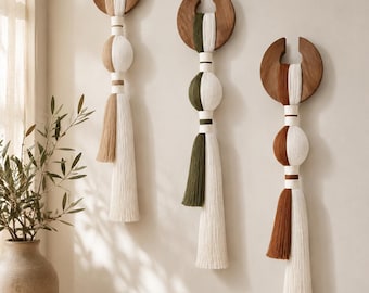 Puede incluir: Tres tapices de pared con piezas superiores de madera oscura en forma de media luna y borlas de fibra vertical en blanco combinadas con beige, verde oliva y marrón terracota. Cada pieza incluye un arco de madera, cordones y flecos.