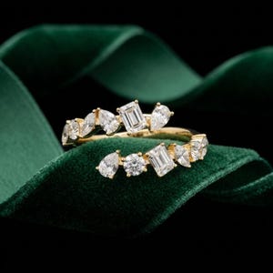 Puede incluir: Un anillo abierto de oro con una serie de piedras transparentes facetadas en varios cortes, incluyendo esmeralda, pera y redondos. Las piedras están dispuestas a lo largo de los dos extremos abiertos de la banda de oro.