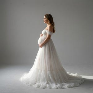 Peut inclure: Une robe de maternité blanche cassé fluide avec un col bateau. La robe présente une jupe à plusieurs épaisseurs et une longue traîne. La personne enceinte se tient le ventre devant un fond gris clair uni.