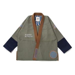May include: Olive green kimono-style jacket with brown trim and navy blue sleeves. The jacket features the text "ALBINO & PRETO" and wave-like designs in blue and white. A circular patch with a similar design is on the lower right.
