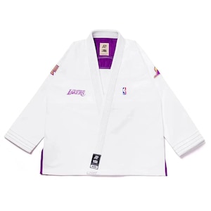 Op de afbeelding: Wit vechtsportuniform met paarse randen en accenten. Het logo van het Los Angeles Lakers-team is paars op de borst weergegeven. Het NBA-logo is ook zichtbaar. Het uniform heeft lange mouwen en een V-hals.