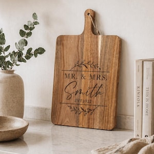 Könnte beinhalten: Ein rechteckiges Holzschneidebrett mit Griff und einer Schlaufe aus Juteschnur. Das Brett ist mit einer Lasergravur versehen, die den Text MR. & MRS. Smith EST2022 zeigt, eingerahmt von zwei horizontalen Linien und dekorativen Blattzweigen.