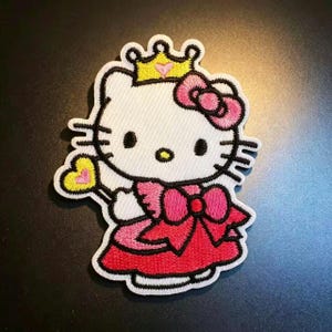 May include: An embroidered iron-on patch featuring a white cartoon cat character wearing a yellow crown with a pink heart, a pink bow on its ear, and a red dress with a large pink bow. The character holds a yellow heart-shaped wand with a pink center.