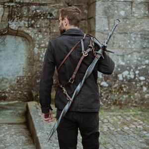 Peut inclure: Une grande épée argentée avec une lame torsadée est portée sur l'épaule avec un harnais en cuir marron. L'épée a une garde et un anneau en haut de la poignée. La personne porte une veste et un pantalon noirs.