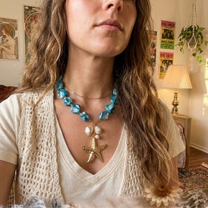 Op de afbeelding: Een turquoise en gouden ketting met een zeesterhanger. De ketting heeft grote, onregelmatige turquoise kralen en een fijne gouden ketting. Een gouden zeesterhanger hangt onder twee witte, parelachtige kralen. De ketting wordt gedragen over een wit shirt.