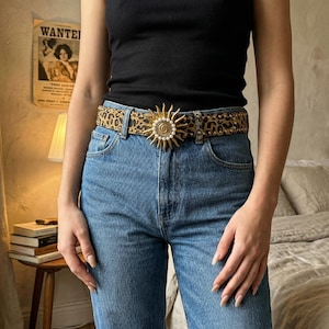 Puede incluir: Un cinturón con estampado de leopardo con una hebilla dorada en forma de sol. El cinturón se lleva con vaqueros azules y una camiseta sin mangas negra. La hebilla del sol tiene una perla en el centro y está rodeada de pequeñas perlas.