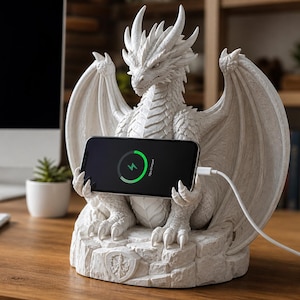 Könnte beinhalten: Ein weißer, drachenförmiger Telefon-Ladeständer. Der Drache hält ein schwarzes Smartphone mit einem Ladesymbol. Der Ständer steht auf einem Holzschreibtisch, im Hintergrund ein Computer und eine kleine Pflanze. Der Drache hat detaillierte Schuppen und Flügel.