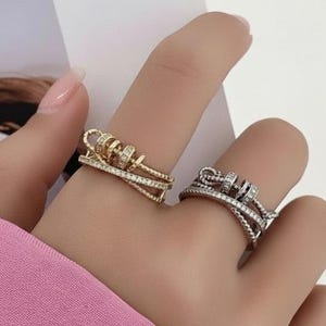 Op de afbeelding: Twee ringen met een beweegbaar abacus-ontwerp met kleine kralen. De ene ring is goudkleurig en de andere zilverkleurig, beide ingelegd met kleine heldere steentjes langs de ringbanden en de centrale horizontale stang van het abacusmechanisme.