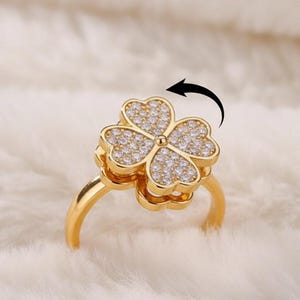 Könnte beinhalten: Ein goldfarbener Ring mit einem vierblättrigen Kleeblatt-Design. Die Kleeblatt-Blütenblätter sind mit kleinen, funkelnden Steinen besetzt. Der Ring ist vor einem weichen, weißen Hintergrund platziert, was sein elegantes Design hervorhebt.