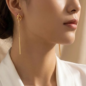 May include: A pair of gold-toned drop earrings featuring a small knot design at the top, connected to a thin, straight metal bar that hangs vertically. The earrings are polished and have a smooth, reflective surface, catching the light.