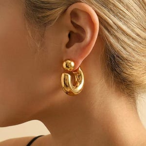 May include: A gold-toned earring featuring a smooth, polished finish. The design consists of a small spherical stud at the top, connected to a thick, curved, C-shaped hoop that hangs below the earlobe, creating a bold and reflective metallic look.
