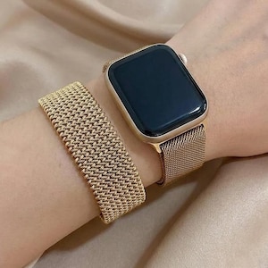 May include: A gold-toned smartwatch with a black screen and a fine mesh metal strap, accompanied by a separate gold-toned woven metal bracelet. The watch has a rectangular case with rounded corners and a matching metallic finish on the strap.