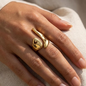 Könnte beinhalten: Ein goldfarbener offener Ring mit zwei markanten, glatten Elementen in organischer Form. Eine Seite des Rings ist tropfenförmig abgerundet, während die andere Seite eine längliche, geschwungene Form aufweist, die sich entlang des Fingers erstreckt.