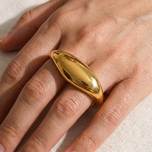 Könnte beinhalten: Ein polierter goldfarbener Ring mit einer glatten, länglichen und gewölbten Oberfläche, die sich über den Finger erstreckt. Das Metall hat ein hochglänzendes, reflektierendes Finish mit einem geschwungenen, massiven Ringdesign.