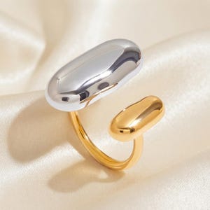 May include: An open-style ring featuring two smooth, rounded, pill-shaped elements. One larger element is finished in a polished silver tone, while the smaller element and the band are finished in a polished gold tone, creating a two-tone design.