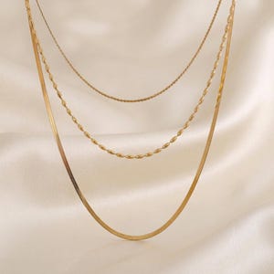 Puede incluir: Un collar en capas de tono dorado que consta de tres cadenas distintas. La cadena superior es un eslabón fino y delicado, la cadena intermedia presenta pequeñas cuentas ovaladas y la cadena inferior es una cadena plana y flexible.
