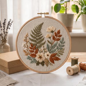 Könnte beinhalten: Ein runder Stickrahmen aus Holz mit einem hellbeigen Stoff, der ein handgesticktes botanisches Motiv zeigt. Die Stickerei besteht aus einer zentralen Anordnung von grünen Farnblättern, rostfarbenem Laub und kleinen cremefarbenen Blüten.