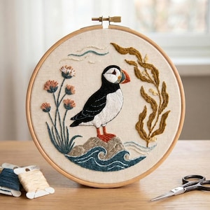 Op de afbeelding: Een ronde houten borduurring met een stoffen paneel waarop een handgeborduurde papegaaiduiker op een rots staat. Het ontwerp bevat oranje, zwart en wit borduurgaren, omringd door zeewier, blauwe golven en bloemstengels.