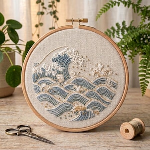May include: A round embroidery hoop featuring a detailed wave design. The wave is depicted in shades of blue and white thread, with a beige background. The wooden hoop is set on a wooden surface, with scissors and thread spools nearby. The craft project is a decorative piece.