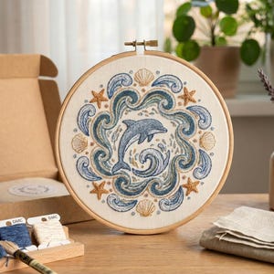 May include: A circular wooden embroidery hoop containing a fabric panel with a hand-stitched design of a central dolphin surrounded by swirling blue and green ocean waves, tan seashells, and orange starfish on an off-white background.