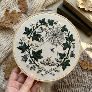 May include: A circular embroidery hoop containing a cream-colored fabric with a hand-stitched design of a spider web, a spider, two moths, three beetles, white flowers, dark green leaves, and two crossed bones at the bottom of the wreath.