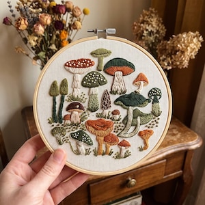 May include: A circular wooden embroidery hoop containing a white fabric panel with detailed thread-stitched illustrations of various mushrooms in shades of green, orange, brown, and white, featuring diverse cap shapes, textures, and stem patterns.