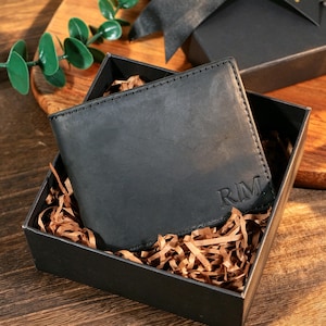 Könnte beinhalten: Ein schwarzes Lederportemonnaie mit den Initialen "RIM" auf der Vorderseite, präsentiert in einer schwarzen Geschenkbox mit geschreddertem Papier. Das Portemonnaie ist rechteckig und scheint neu zu sein. Die Box ist geöffnet und zeigt das Portemonnaie und das Verpackungsmaterial.