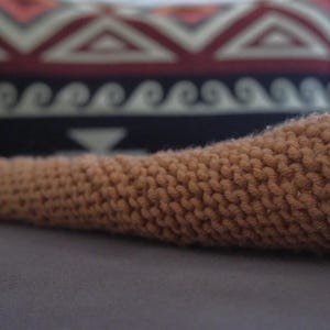 Könnte beinhalten: Nahaufnahme eines braunen Strickartikels, möglicherweise ein Glied eines Stofftiers oder ein dekoratives Element. Das Gestrick hat ein strukturiertes Aussehen. Der Hintergrund ist verschwommen und zeigt einen gemusterten Stoff mit geometrischen und wellenartigen Designs.