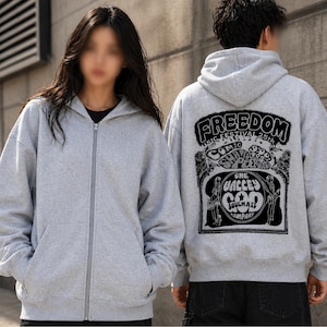Könnte beinhalten: Zwei graue Kapuzenpullover mit Reißverschluss. Der Kapuzenpullover auf der Vorderseite hat einen durchgehenden Reißverschluss und Taschen. Der Kapuzenpullover auf der Rückseite hat eine schwarze Grafik mit den Worten "FREEDOM MUSIC FESTIVAL 25TH" und anderem Text. Die Kapuzenpullover sind lässig.