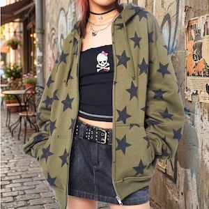 May include: Olive green zip-up hoodie with a black star pattern. The hoodie has a drawstring hood and front pockets. The person is wearing a black tube top with a skull graphic and a black denim skirt.