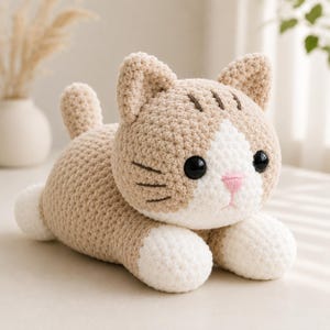 Può includere: Un peluche a forma di gatto realizzato all'uncinetto con una combinazione di colori marrone chiaro e bianco. Il gatto ha grandi occhi in plastica nera, un piccolo naso ricamato in rosa e baffi ricamati marrone scuro. È raffigurato sdraiato con zampe bianche.