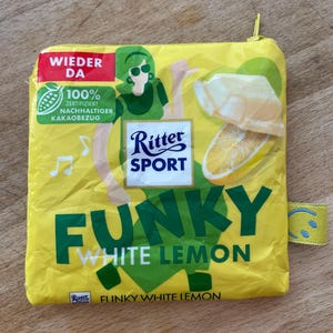 Könnte beinhalten: Eine gelbe Reißverschlusstasche mit dem Design der Ritter Sport Funky White Lemon Schokoladenverpackung. Die Vorderseite zeigt eine Grafik einer Person, ein Stück weiße Schokolade, eine Zitronenscheibe sowie den Text WIEDER DA, 100% ZERTIFIZIERT NACHHALTIGER KAKAO BEZUG, Ritter Sport und FUNKY WHITE LEMON.